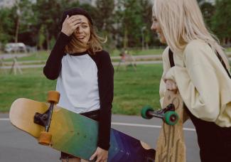 A teenager holding a yellow and blue longboard talking to her friend